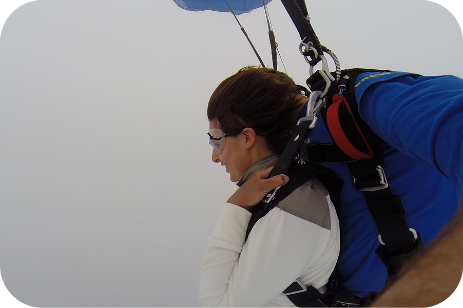 Sara Rosenblatt mit Schutzbrille im Tandemgurt beim Fallschirmsprung über Sydney, darüber der geöffnete Schirm