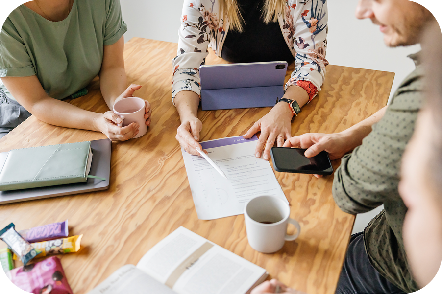 Drei Personen sitzen an einem Holztisch und besprechen beim Auftaktmeeting Unterlagen, daneben Tablet, Notizbuch und Kaffeetassen