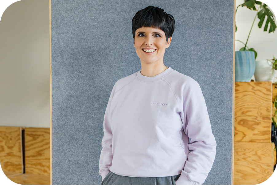 Sara Rosenblatt lächelt in einem hellen Büro vor einer grauen Filzwand, im Hintergrund Holzmöbel und eine Pflanze