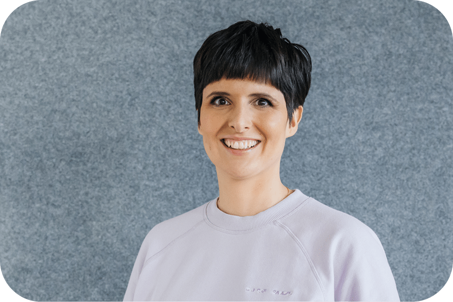 Sara Rosenblatt lächelt in einem hellen Büro vor einer grauen Filzwand, im Hintergrund Holzmöbel und eine Pflanze